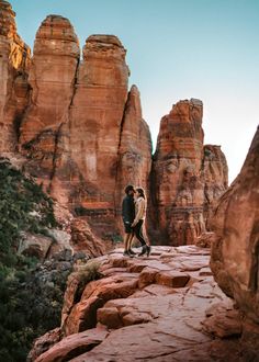 two people standing on the edge of a rock formation in front of some red rocks