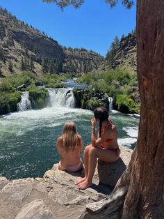 two women are sitting on the rocks near a waterfall and looking at something in the distance