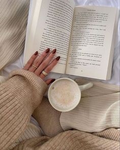 a woman laying in bed reading a book and holding a cup of coffee next to her