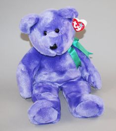 a purple teddy bear with a green ribbon on it's neck sitting against a gray background