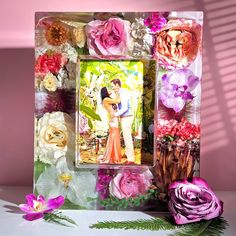 a photo frame with flowers on it and a flower arrangement next to it, in front of a pink wall