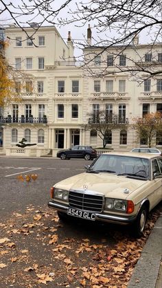 an old mercedes parked in front of a large white building with autumn leaves on the ground