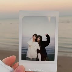 two women standing next to each other on the beach holding up polaroids with their hands