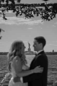 A couple embraces in the foreground, while the Statue of Liberty stands prominently across the water in the background. The image is rendered in black and white. The Statue Of Liberty, Skyline View, Brooklyn Wedding, Monochrome Photography