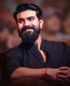 a man with a beard and black shirt sitting in front of a brown wall smiling