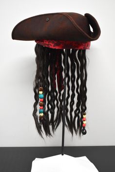 Complete your Pirate Costume this season with this Deluxe Caribbean Pirate Hat with Attached Dread Locs Wig. Perfect for Halloween, Cosplay, Theater and more! Featuring a tri-corne hat with attached brown faux braided hair with beads accented by a red band with floral filigree. Great for Men or Women! Tricorner hat has sweatband inside for a customized fit. Fits approximately 51 cm to 58 cm. Dreads hand approximately 15" long. Cap stands approximately 5" tall. Handmade, assembled, and crafted by hand with assistance from hand driven fabric forming and hand driven sewing equipment. *partially handmade, partially machine sewn.* Adjustable Themed Costume Accessories For Cosplay Events, Themed Adjustable Costume Hats For Cosplay, Adjustable Pirate Costume Accessories For Costume Party, Pirate Costume Accessories For Costume Party, Adjustable Pirate Costume Accessories For Halloween, Straw Pirate Hat, Tricorn Pirate Hat Diy, Authentic Pirate Hat, Brown Pirate Hat