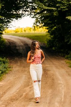 a woman walking down a dirt road with trees in the background