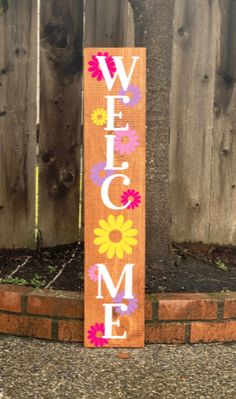 a welcome sign with flowers on it in front of a wooden fence and brick wall