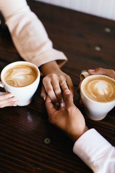 two people holding hands while sitting at a table with cups of coffee in front of them