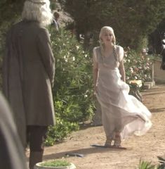 a woman in a white dress walking down a dirt road next to trees and bushes