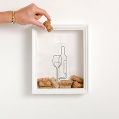 a person holding a wine glass and corks in front of a white frame with a drawing on it