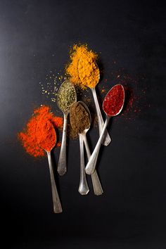 five spoons filled with different colored spices on top of a black surface, next to each other