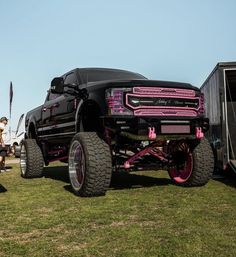 a large black truck with pink wheels parked in the grass next to other trucks and people