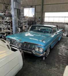 an old blue car is parked in a garage with other cars and trucks behind it