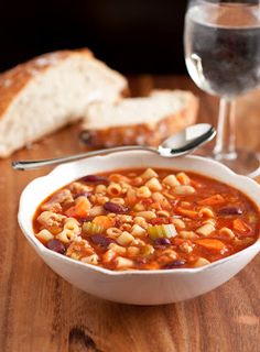 a bowl of soup with beans and carrots on a wooden table next to a glass of wine