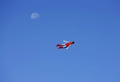 a red and white plane flying in the sky with a half moon behind it on a clear day