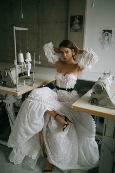 a woman sitting at a sewing machine wearing a white dress with ruffles on it