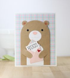 a brown bear holding a pink envelope with the word hugs on it's chest