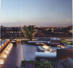 an outdoor deck with seating and lights on it at night in front of the cityscape