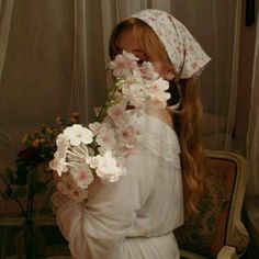 a woman with long hair wearing a white dress and holding flowers in her hand while sitting on a chair