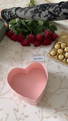 a heart shaped box filled with chocolates next to red roses and a woman's hand