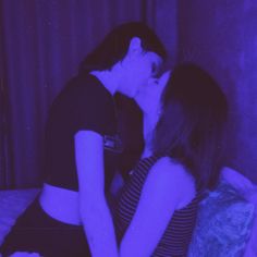 two women sitting on a bed in front of a purple light and one is kissing the other