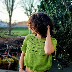 a woman wearing a green crochet top talking on her cell phone while standing next to a tree