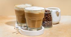 two glasses filled with liquid sitting on top of a table next to coffee beans and a teapot