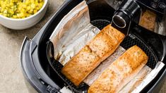 two fish fillets in an air fryer next to a bowl of corn on the cob