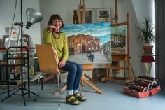 a woman is sitting on a chair in front of an easel with paintings behind her