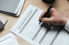 a person filling out a college application form on a desk with other papers and laptops