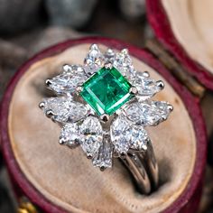 an emerald and diamond ring sitting on top of a velvet case in a wooden box