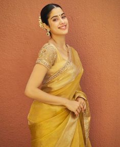 The image showcases a Janhvi Kapoor  wearing a stunning mustard-yellow sari with a golden sheen. The sari features a subtle, intricate border that enhances its luxurious feel, while the blouse is richly embroidered with detailed work that adds texture and elegance. The overall look is traditional yet sophisticated, with a neatly draped sari and matching accessories. Her hair is styled in a sleek bun adorned with jasmine flowers, contributing to the classic look. The addition of statement earrings and a bindi completes the ensemble, exuding grace and timeless beauty. The simple, earthy-toned background highlights the vividness of the sari and the warmth of the look. भारतीय दुल्हन संबंधी, डिजाइनर कपड़े, Golden Saree, Janhvi Kapoor