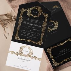 two black and gold wedding stationery cards on top of a table with dried flowers