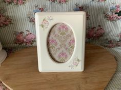 a white and pink flowered photo frame on top of a wooden table next to wallpaper