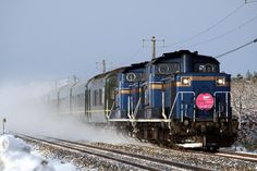 a blue train traveling down tracks next to snow covered ground and power lines in the background