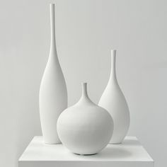 three white vases sitting on top of a white shelf in front of a wall