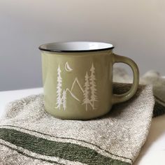 a green coffee mug sitting on top of a towel next to a cup with trees drawn on it