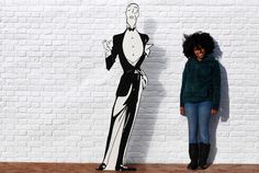 a woman standing next to a drawing of a man in a tuxedo on a white brick wall