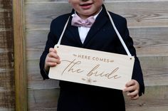 a little boy in a tuxedo holding a sign that says here comes the bride