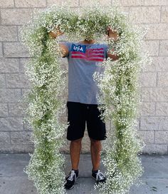 a man standing in front of a wall covered in white flowers and greenery with his hands behind his head