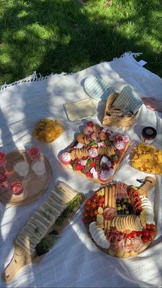 a picnic table with food and drinks on it in the shade, including breads, crackers, cheeses, fruit, and vegetables