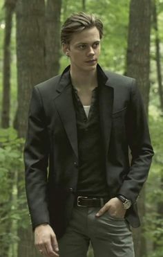 a young man standing in the woods wearing a black suit and tie with his hands in his pockets