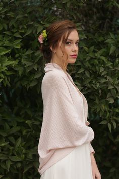 a woman standing in front of some bushes wearing a white dress and pink cardigan