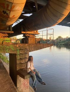 a woman sitting on the edge of a concrete wall next to water under an overpass