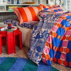 a bed with colorful sheets and pillows on top of it next to a red table