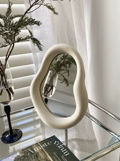 a glass table with a book and mirror on it next to a plant in a vase