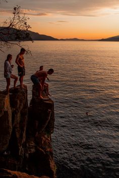 three boys are standing on the edge of a cliff by the water at sunset or dawn