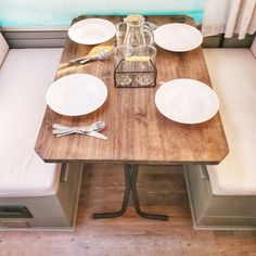 a wooden table with white plates and silverware on it, in front of a window