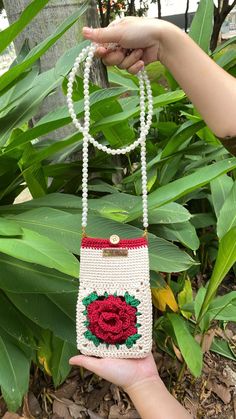 a hand holding a white purse with a red rose on the front and green leaves behind it